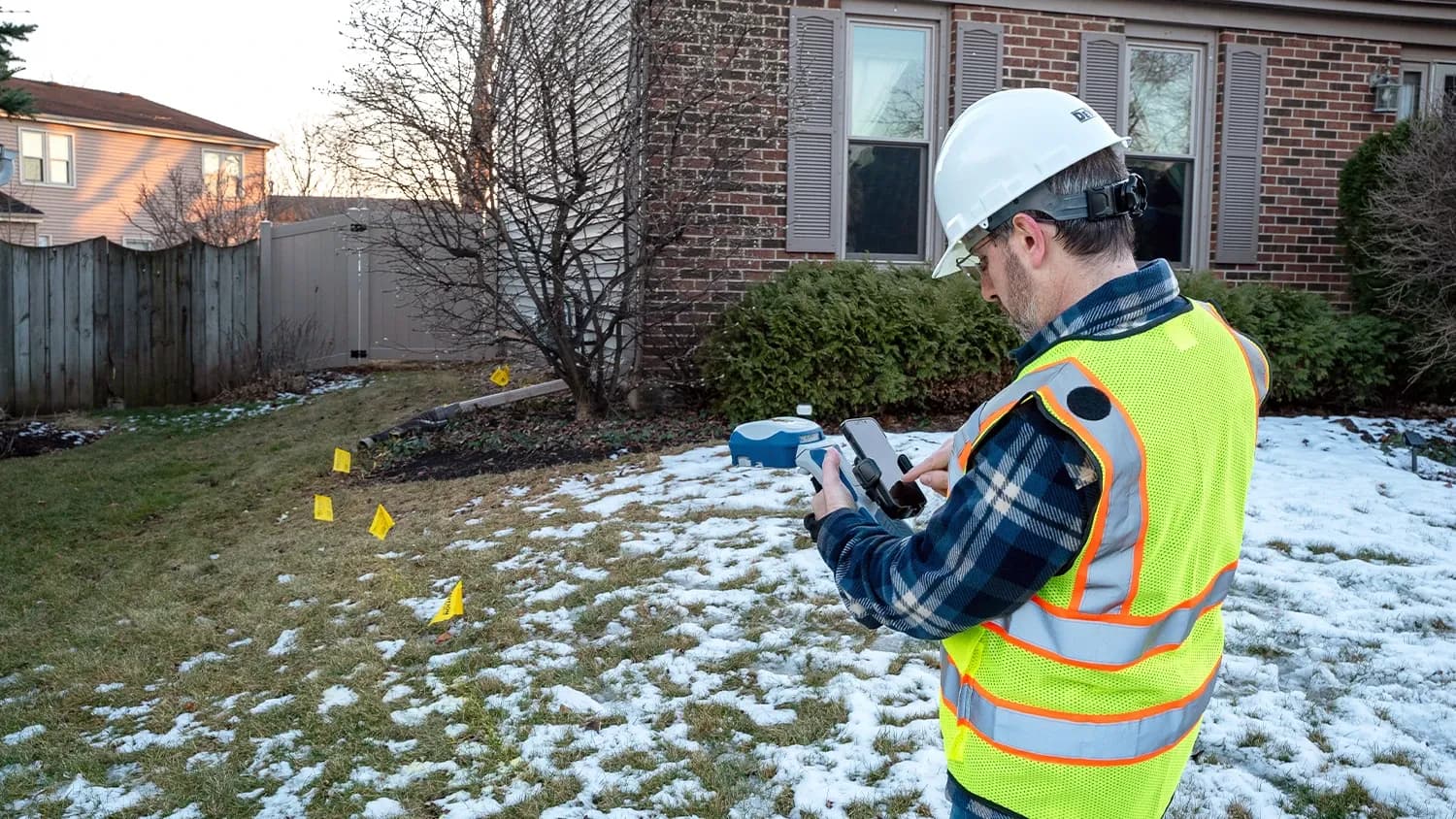 Utility technician staking a gas line with a handheld GNSS receiver, locate flags marked in the snow